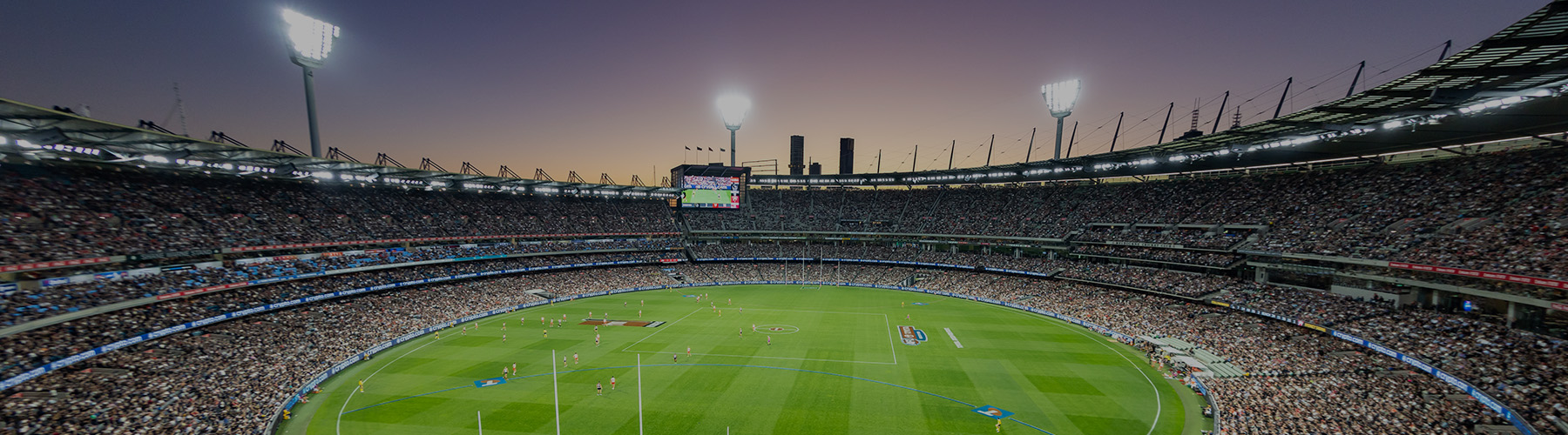 A wide image of the MCG stadium under lights with the sunset in the background