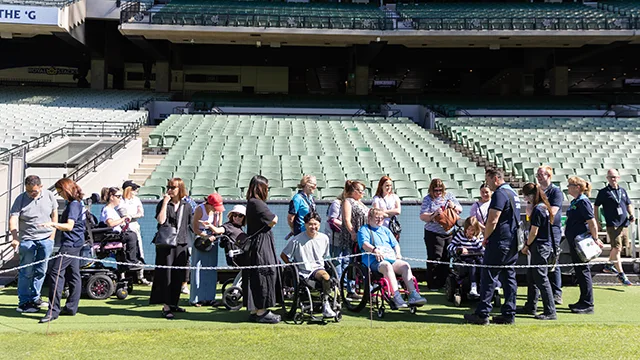 A group tours an empty stadium along the boundary line with some group members using wheelchairs and mobility aides.