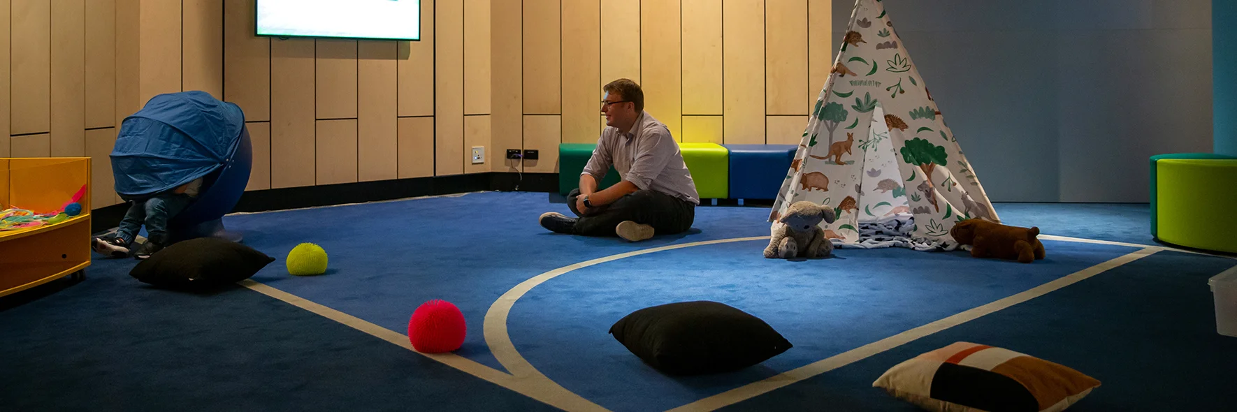 Guardian sits near child using tent in a sensory space. The room is open-plan, with soft furniture and fidgets.