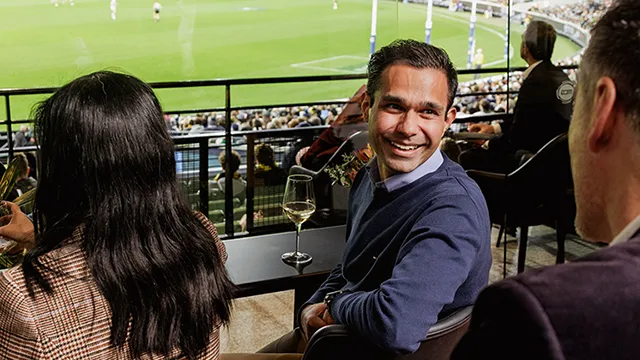 Happy guests enjoying MCG AFL game in private lounge area with wine and goal view.
