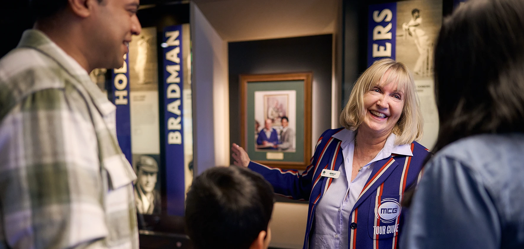 A smiling MCG tour guide shows a family a display