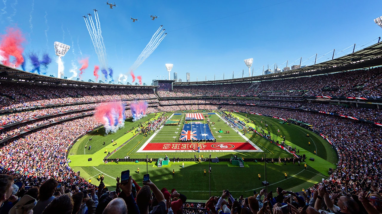 Packed MCG crowd watching a pre-game NFL ceremony, with US and Australian flags on the field and a jet flyover.